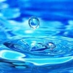Uma gota de água cristalina suspensa no ar acima da superfície azul da água, criando ondas circulares ao cair.