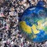 Representação da poluição e impactos ambientais mostrada por um globo terrestre parcialmente coberto por uma grande quantidade de lixo e resíduos espalhados ao redor.